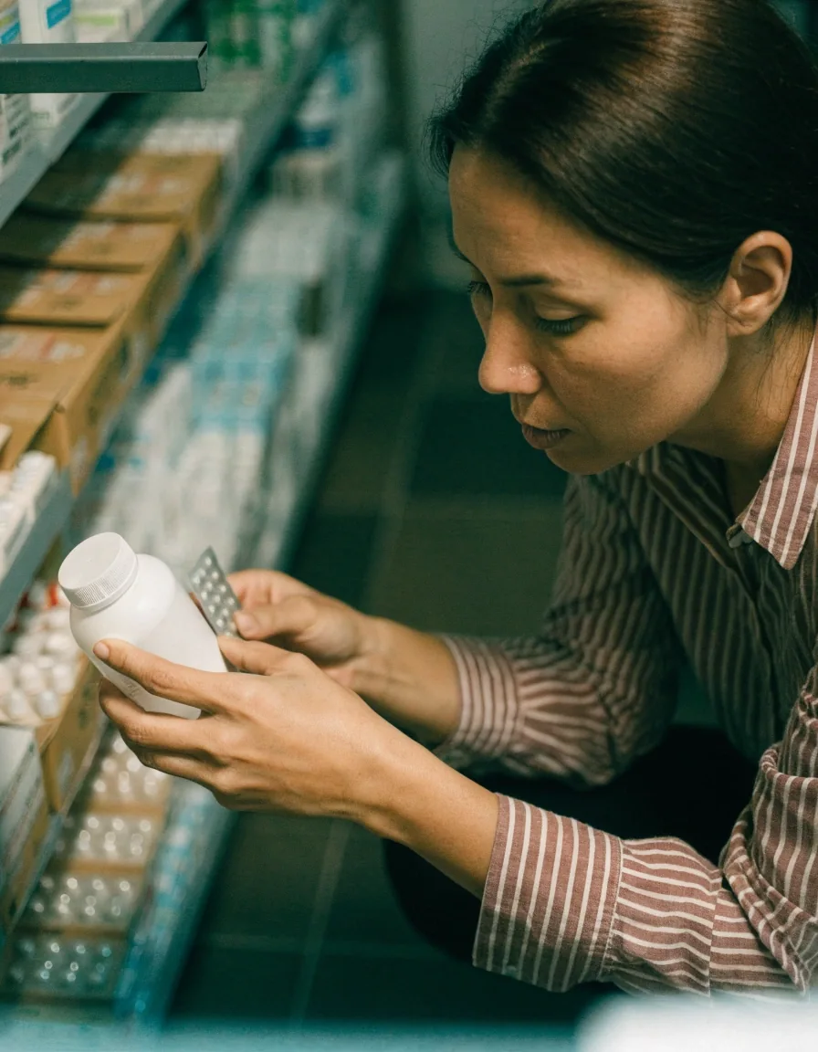 An unmarked white bottle standing on a pharmacy shelf beside a stack of unlabelled blister cards and a saucer of mixed round tablets